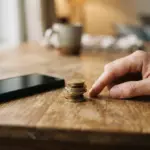 A stack of coins sits on a wooden table next to a smartphone and a hand