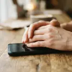 Hands resting on a smartphone placed on a wooden table