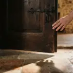 A close-up of a hand pausing at the threshold of a sturdy, slightly ajar dark wooden door